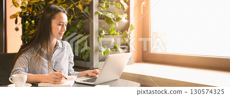 A woman sits at a table in a cafe, focused on her laptop while taking notes, surrounded by greenery and natural light, copy space A woman sits at a table in a cafe, focused on her laptop while taking notes, surrounded by greenery and natural light, copy space 130574325