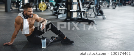 A young man sits on the gym floor, taking a break after exercising, with a towel around his neck and a water bottle nearby, copy space A young man sits on the gym floor, taking a break after exercising, with a towel around his neck and a water bottle nearby, copy space 130574461