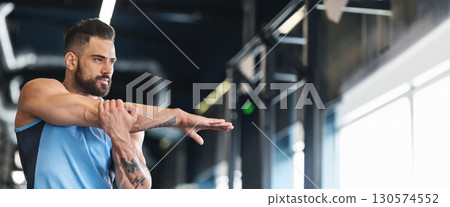 A man performs arm stretches in a modern gym, focusing on flexibility and fitness as sunlight streams through large windows, copy space 130574552