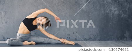A young woman is performing a side stretch while practicing yoga on a mat in a minimalist indoor space, copy space 130574621