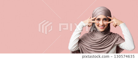 Playful arabic girl in hijab showing v-sign symbols near eyes, having fun over pink background with free space, closeup Playful arabic girl in hijab showing v-sign symbols near eyes, having fun over pink background with free space, closeup 130574635