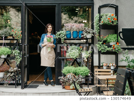 Startup, small business, eco restaurant outdoor and modern rustic flower shop. Smiling millennial beautiful female in apron holds bouquet of tulips for client, at front door of plants studio or cafe 130574649
