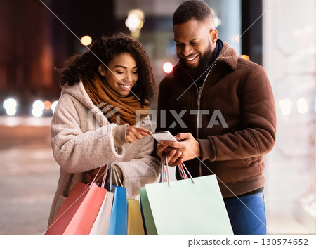 App For Online Shopping. Portrait of smiling black man and woman using mobile phone, lady pointing at screen, couple holding shopper bags, standing outdoors in the evening, choosing gift 130574652