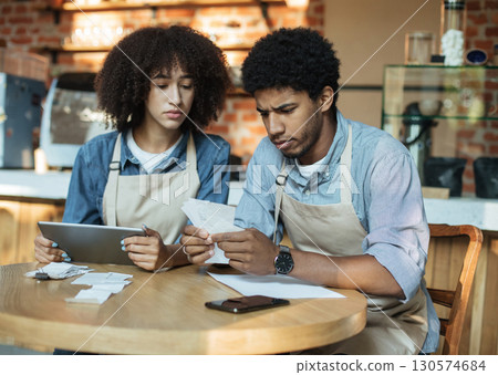 Problems in small business during covid-19 lockdown. Sad millennial african american couple owners in aprons work with accounts, and solve debt problems bookkeeping in cafe, empty space Problems in small business during covid-19 lockdown. Sad millennial african american couple owners in aprons work with accounts, and solve debt problems bookkeeping in cafe, empty space 130574684