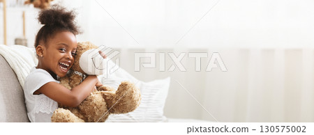 A young black girl with curly hair joyfully hugs a fluffy teddy bear while seated comfortably on a couch. She is smiling widely, radiating happiness in a cozy indoor setting, copy space A young black girl with curly hair joyfully hugs a fluffy teddy bear while seated comfortably on a couch. She is smiling widely, radiating happiness in a cozy indoor setting, copy space 130575002
