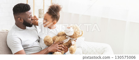 African American father and his daughter share a delightful moment together in their cozy living room. The little girl holds a teddy bear while smiling at her dad, copy space 130575006