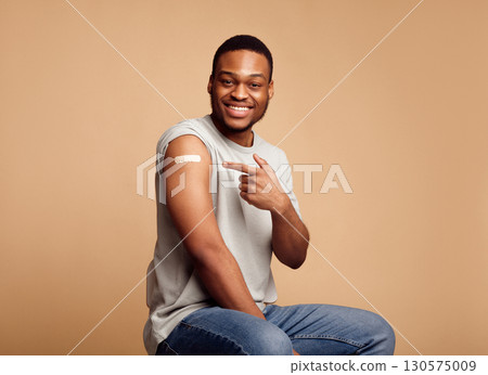 Vaccination Against Covid-19. Joyful Vaccinated African Guy Pointing Finger At Adhesive Plaster Bandage On His Arm After Coronavirus Antiviral Injection Sitting On Beige Background. I've Got Vaccine Vaccination Against Covid-19. Joyful Vaccinated African Guy Pointing Finger At Adhesive Plaster Bandage On His Arm After Coronavirus Antiviral Injection Sitting On Beige Background. I've Got Vaccine 130575009
