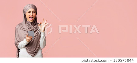 Cool islamic app. Positive muslim girl in hijab winking and showing ok gesture while using smartphone, standing over pink background with copy space Cool islamic app. Positive muslim girl in hijab winking and showing ok gesture while using smartphone, standing over pink background with copy space 130575044