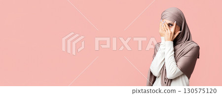 Modesty concept. Shy muslim girl in hijab peeking through fingers with interest, posing over pink background with empty space Modesty concept. Shy muslim girl in hijab peeking through fingers with interest, posing over pink background with empty space 130575120