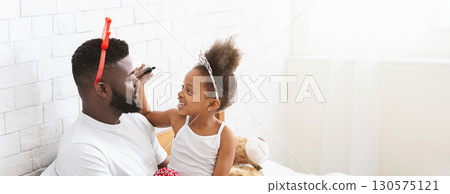 Black father and his daughter are having a joyful moment in a brightly lit bedroom. The little girl, wearing a crown, is playfully applying makeup to her dad's face while sitting on the bed 130575121