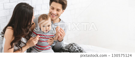 A joyful family is enjoying quality time together in their bedroom. The mother and father are playfully interacting with their young child, who is smiling and engaging with them, copy space 130575124