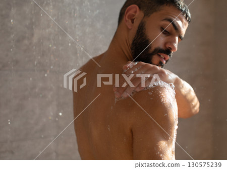 Skincare Concept. Young muscular man taking refreshing shower washing his body, back and bare shoulders with soap foam. Confident handsome guy standing in the bathroom under falling hot water drops 130575239