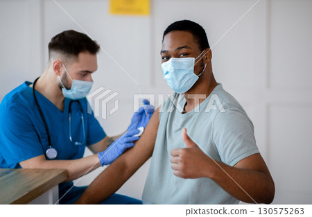 African American man in antiviral mask gesturing thumb up during coronavirus vaccination, approving of covid-19 immunization. Young doctor giving vaccine injection to male patient African American man in antiviral mask gesturing thumb up during coronavirus vaccination, approving of covid-19 immunization. Young doctor giving vaccine injection to male patient 130575263