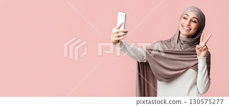 Peace selfie. Cheerful arabic girl in hijab taking self-portrait on smartphone and showing v-sign over pink background with free space Peace selfie. Cheerful arabic girl in hijab taking self-portrait on smartphone and showing v-sign over pink background with free space 130575277