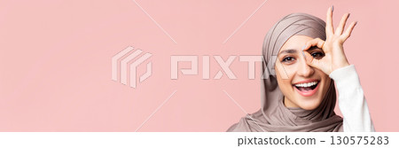 Cheerful arabic lady in hijab showing ok gesture, holding hand near eye, looking through fingers at camera, pink background with empty space, closeup 130575283