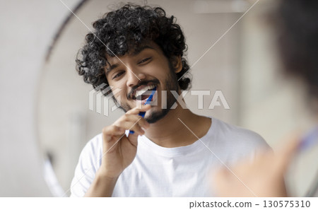 Oral care concept. Young indian man cleaning teeth with toothbrush, smiling to his reflection in mirror, doing toothcare hygiene routine in the morning in bathroom 130575310