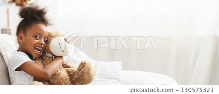 African American young girl with curly hair looks delighted as she hugs a soft teddy bear in a bright, cozy living room. Sunlight filters through sheer curtains, creating a warm atmosphere. African American young girl with curly hair looks delighted as she hugs a soft teddy bear in a bright, cozy living room. Sunlight filters through sheer curtains, creating a warm atmosphere. 130575321