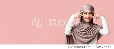 Mysterious muslim girl in hijab holding two fingers near eyes, showing v-sign symbols, playfully framing her glance 130575337