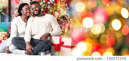 We wish you a merry Christmas. Portrait of happy afro couple sitting on floor near Xmas tree, celebrating winter holidays at home 130575339