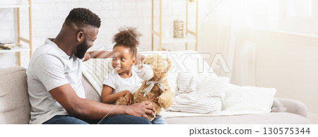 Black father and his young daughter share a joyful moment while sitting on a comfortable couch at home. The girl holds a teddy bear, smiling and engaging with her dad, copy space Black father and his young daughter share a joyful moment while sitting on a comfortable couch at home. The girl holds a teddy bear, smiling and engaging with her dad, copy space 130575344
