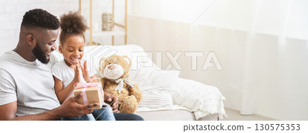 Black father and daughter sit closely on a cozy couch, eagerly unwrapping a gift. The little girl, filled with excitement, claps her hands while her father watches with delight, copy space 130575353