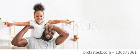 Black father and daughter are joyfully interacting in a well-lit living room, with the girl perched on his shoulders. She smiles widely, arms outstretched, while he supports her playfully, copy space 130575361