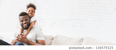 A joyful black father interacts with his young daughter, who playfully hugs him from behind while they sit on a comfortable sofa. The warm atmosphere highlights their close bond, copy space A joyful black father interacts with his young daughter, who playfully hugs him from behind while they sit on a comfortable sofa. The warm atmosphere highlights their close bond, copy space 130575525