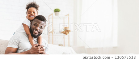 African American father and daughter are enjoying a special bonding moment in their cozy living room. The girl, smiling widely, is playfully hugging her dad from behind while he sits comfortably 130575538