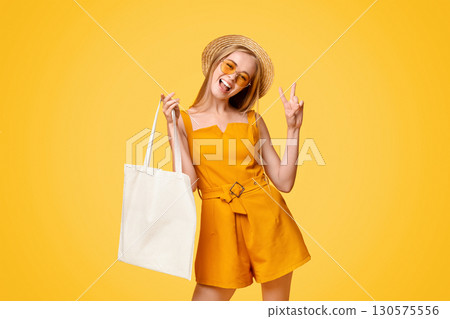 Joyful Teen Girl Holding Eco Tote Bag With Mockup Place For Design And Showing Peace Gesture Over Yellow Background Joyful Teen Girl Holding Eco Tote Bag With Mockup Place For Design And Showing Peace Gesture Over Yellow Background 130575556