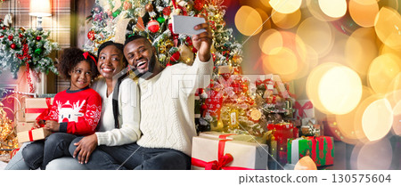 Happy african american family of three taking selfie near Christmas tree at home, sitting on floor close to fireplace in decorated living room Happy african american family of three taking selfie near Christmas tree at home, sitting on floor close to fireplace in decorated living room 130575604