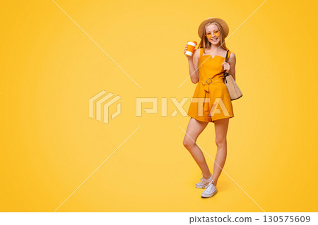 Summer Girl. Stylish Young Woman In Hat. Sunglasses And Jumpsuit Enjoying Her Takeaway Drink Over Yellow Studio Background, Copy Space 130575609