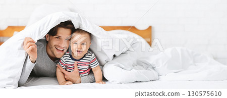A father and his baby share a delightful moment together, both smiling warmly while snuggled under a soft white blanket. They lie comfortably on bed, surrounded by a comforting atmosphere 130575610