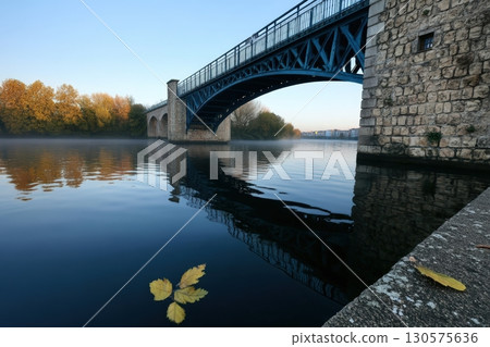 Calm waters reflect an autumn bridge in early morning light near a peaceful riverbank 130575636