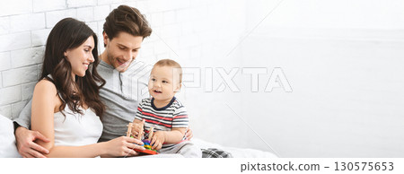 A joyful family engages in playful activities together in a bright living room. The parents sit on a comfortable bed with their toddler, who eagerly plays with colorful toys, copy space A joyful family engages in playful activities together in a bright living room. The parents sit on a comfortable bed with their toddler, who eagerly plays with colorful toys, copy space 130575653