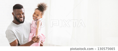A joyful black father holds his smiling daughter in his arms as they dance playfully. Sunlight filters through the bright room, illuminating their happiness and creating a warm atmosphere. A joyful black father holds his smiling daughter in his arms as they dance playfully. Sunlight filters through the bright room, illuminating their happiness and creating a warm atmosphere. 130575665