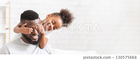 A joyful black father is sitting comfortably while his young daughter playfully covers his eyes with her hands. Their cheerful expressions highlight the loving connection they share, copy space 130575668