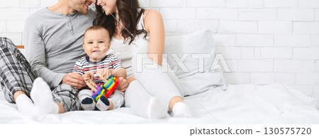A joyful family sits close together on a comfortable bed, with the parents smiling at each other while their young child plays happily. The warm and inviting atmosphere is enhanced by soft bedding 130575720