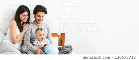 A mother and father sit together on a comfortable bed, actively engaging their young child who is fascinated by a globe. The parents share smiles and joy as they explore the world, copy space 130575736