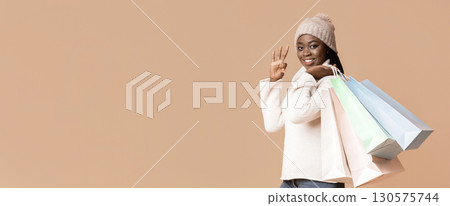 A young black woman smiles brightly while wearing a cozy sweater and a warm hat. She holds colorful shopping bags in one hand, copy space A young black woman smiles brightly while wearing a cozy sweater and a warm hat. She holds colorful shopping bags in one hand, copy space 130575744