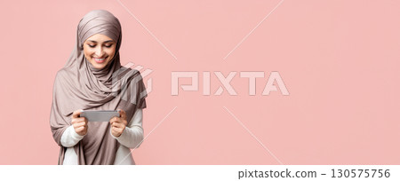 Millennial muslim woman in hijab holding smartphone in horizontal orientation, watching videos or playing games over pink background with empty space Millennial muslim woman in hijab holding smartphone in horizontal orientation, watching videos or playing games over pink background with empty space 130575756