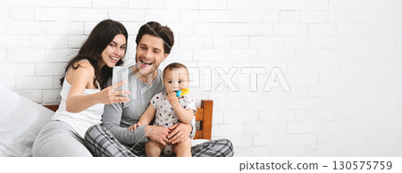 A joyful family enjoys a moment together as they capture memories using a smartphone. The parents are seated on the bed with their baby, sharing smiles and laughter in a bright, copy space A joyful family enjoys a moment together as they capture memories using a smartphone. The parents are seated on the bed with their baby, sharing smiles and laughter in a bright, copy space 130575759