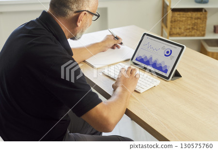 Man reviewing analytics dashboard on tablet and taking notes at home desk Man reviewing analytics dashboard on tablet and taking notes at home desk 130575760