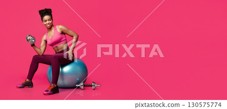Black fitness girl exercising with dumbbells, sitting on fitness ball over red studio background, free space 130575774