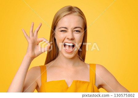 Everything is Great. Beautiful Young Girl Winking And Showing Ok Sign, Standing Over Yellow Background With Copy Space 130575802