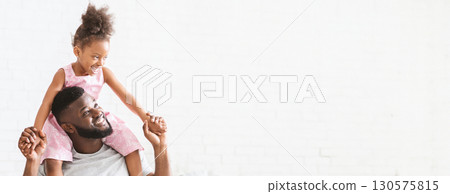A cheerful black father enjoys playful moments with his daughter, who is happily perched on his shoulders. Their laughter fills the air as they engage in a joyful activity, copy space A cheerful black father enjoys playful moments with his daughter, who is happily perched on his shoulders. Their laughter fills the air as they engage in a joyful activity, copy space 130575815