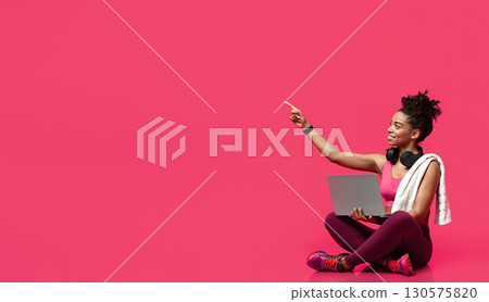 Smiling fitness afro girl with laptop, towel and earphones pointing up at copy space over red background, panorama, sales at sportswear shop 130575820