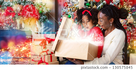 Christmas magic. African american mom and daughter opening shining Christmas gift box, sitting near Xmas tree, celebrating holidays at home 130575845