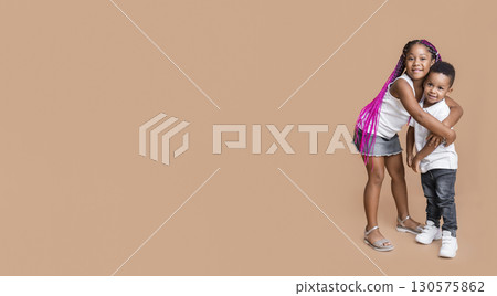 Siblings love. Cute little afro brother and sister cuddling and smiling at camera over yellow studio background, free space 130575862
