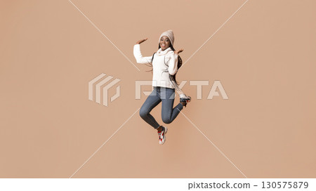 Playful young african woman in winter hat having fun over beige studio background, jumping in the air gracefully 130575879