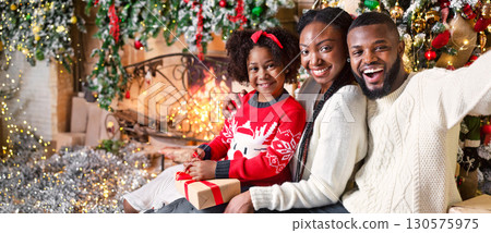 Happy afro parents taking selfie with their cute little daughter near Christmas tree, sitting on floor in decorated living room Happy afro parents taking selfie with their cute little daughter near Christmas tree, sitting on floor in decorated living room 130575975
